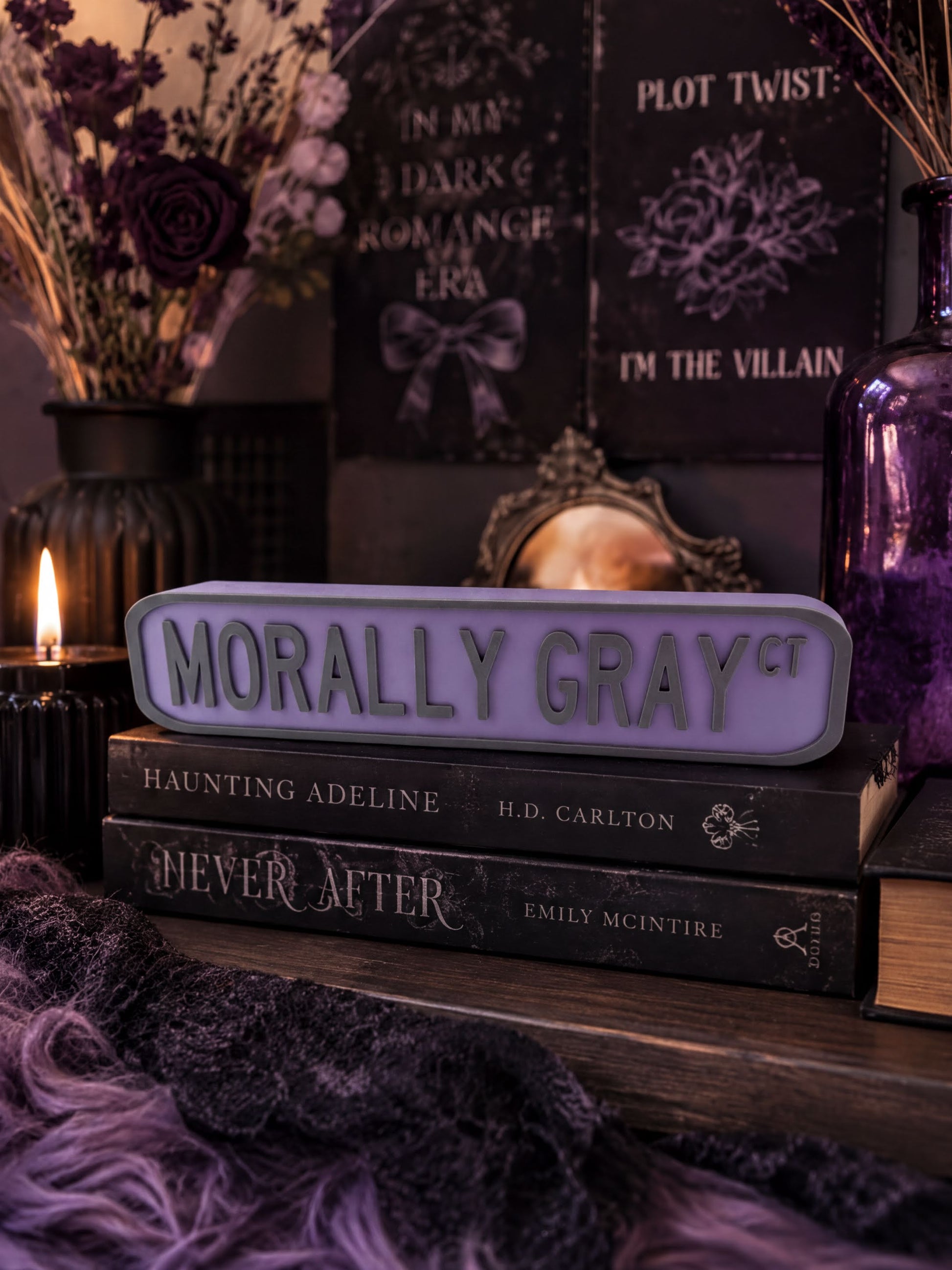 a dark and moody atmosphere with a purple sign that reads "Morally Gray" in the foreground. Behind the sign, there are several books, including "Haunting Adeline" and "Never After". The background includes a vase with purple flowers and a mirror, adding to the overall dark and mysterious ambiance of the scene.
