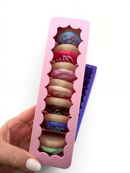 A hand is holding a pink silicone mold with a scalloped edge, which is filled with a variety of colorful donuts.