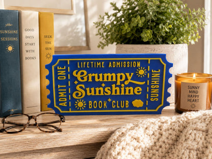 a blue ticket-shaped sign with yellow text, a candle, glasses, and a potted plant on a wooden surface.