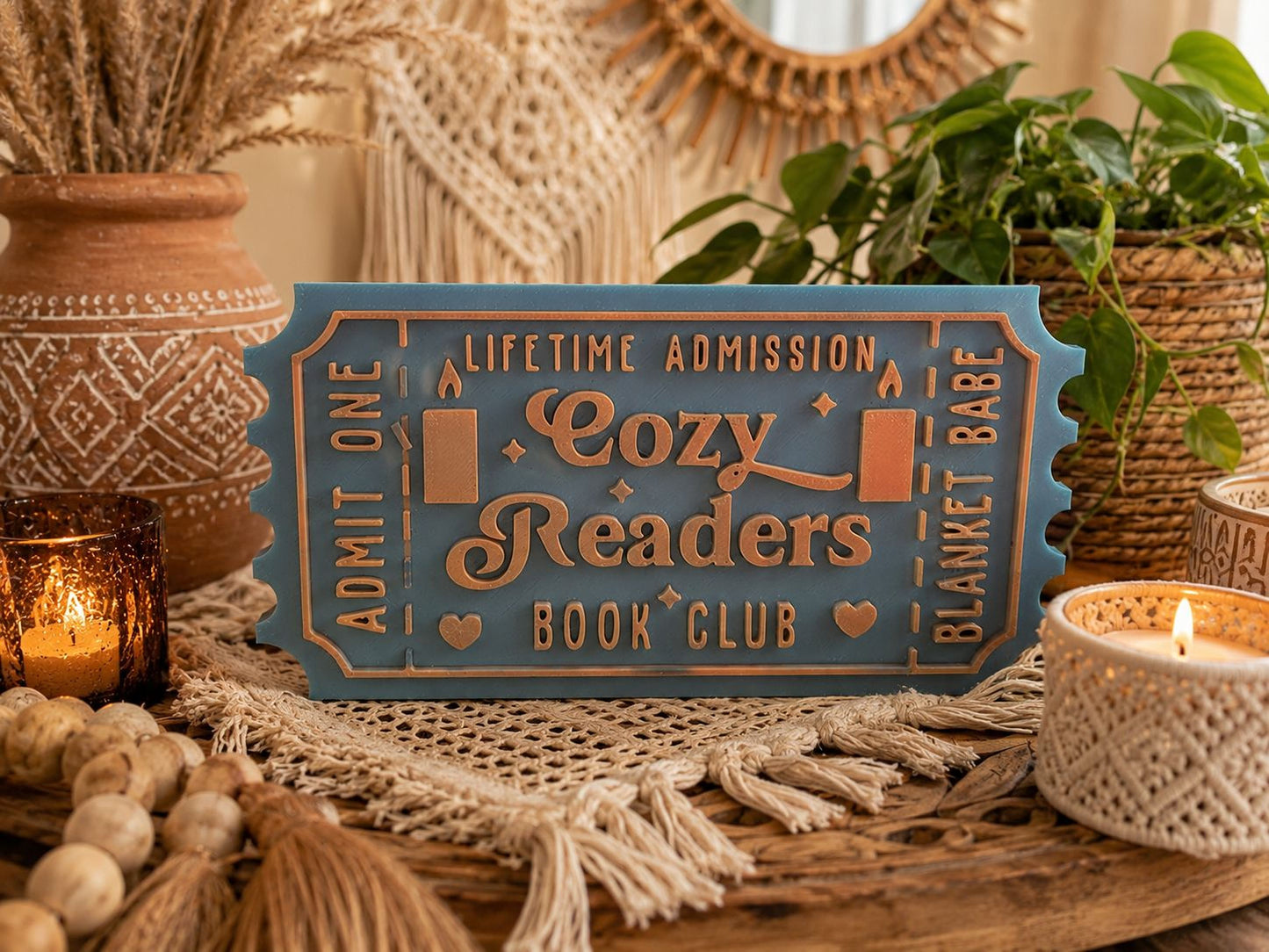A blue ticket-shaped sign with the text "Lifetime Admission Cozy Readers Book Club" is displayed on a wooden surface, surrounded by various decorative items such as plants, candles, and woven baskets.