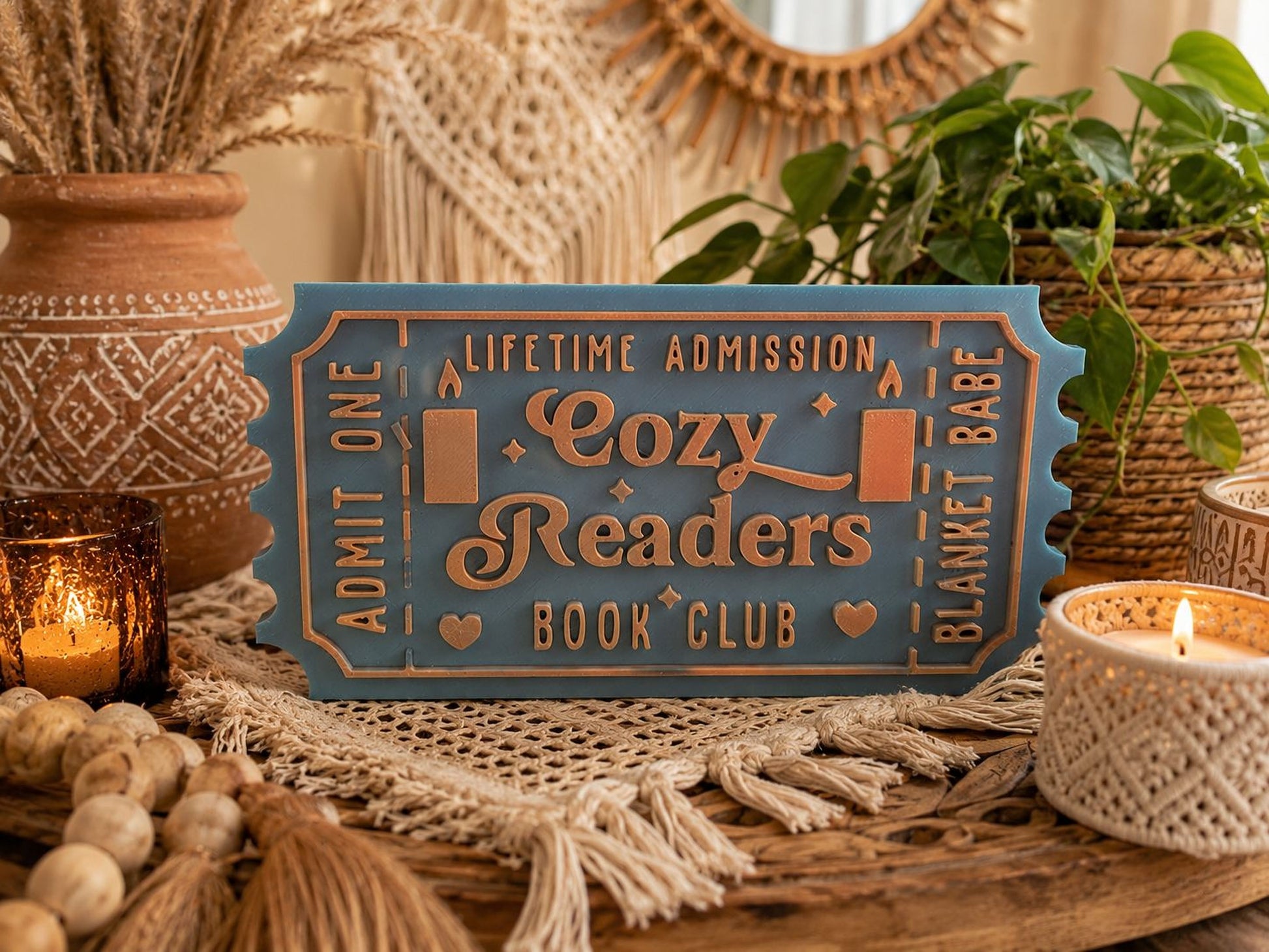 A blue ticket-shaped sign with the text "Lifetime Admission Cozy Readers Book Club" is displayed on a wooden surface, surrounded by various decorative items such as plants, candles, and woven baskets.