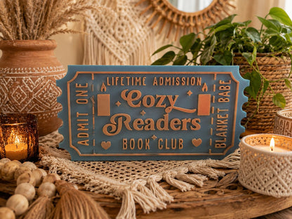 A blue ticket-shaped sign with the text "Lifetime Admission Cozy Readers Book Club" is displayed on a wooden surface, surrounded by various decorative items such as plants, candles, and woven baskets.