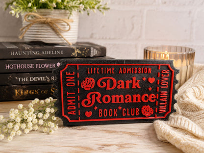 a black ticket with red text, a stack of books, a candle, and some flowers on a table.