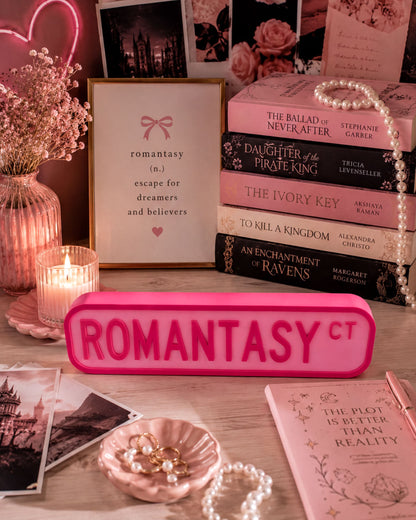 a pink sign with the word "ROMANTASY" in large letters, surrounded by various pink-themed items such as books, flowers, and jewelry.