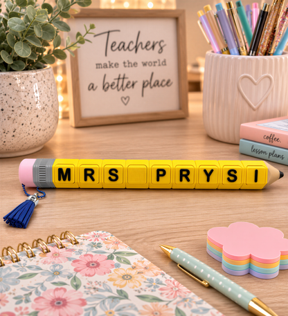 a desk with various school supplies, including a yellow pencil with the letters "MRS PRYS" on it, a notebook, a pen, and a plant. There is also a framed sign that says "Teachers make the world a better place".