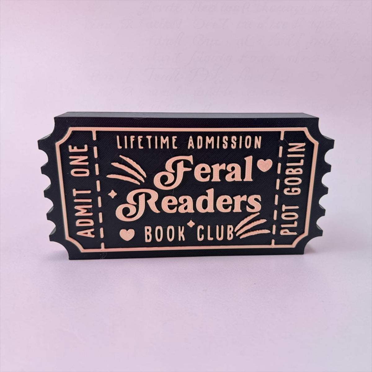 A black and gold ticket-shaped object with text that reads "Lifetime Admission Feral Readers Book Club Plot" printed on it.