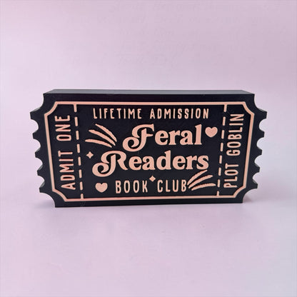 A black and gold ticket-shaped object with text that reads "Lifetime Admission Feral Readers Book Club Plot" printed on it.