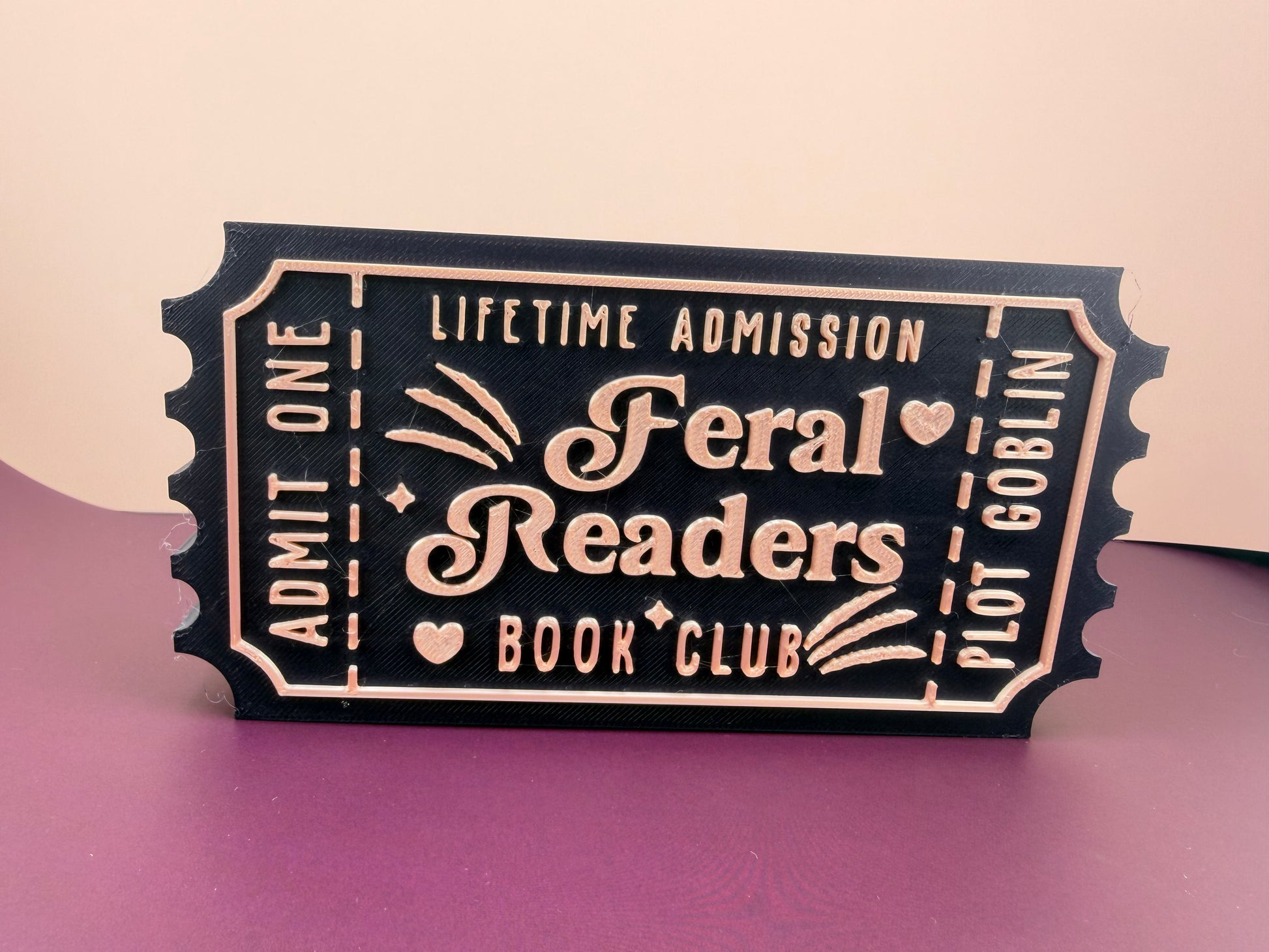A black and gold ticket-shaped sign with the text "Feral Readers Book Club" printed on it.