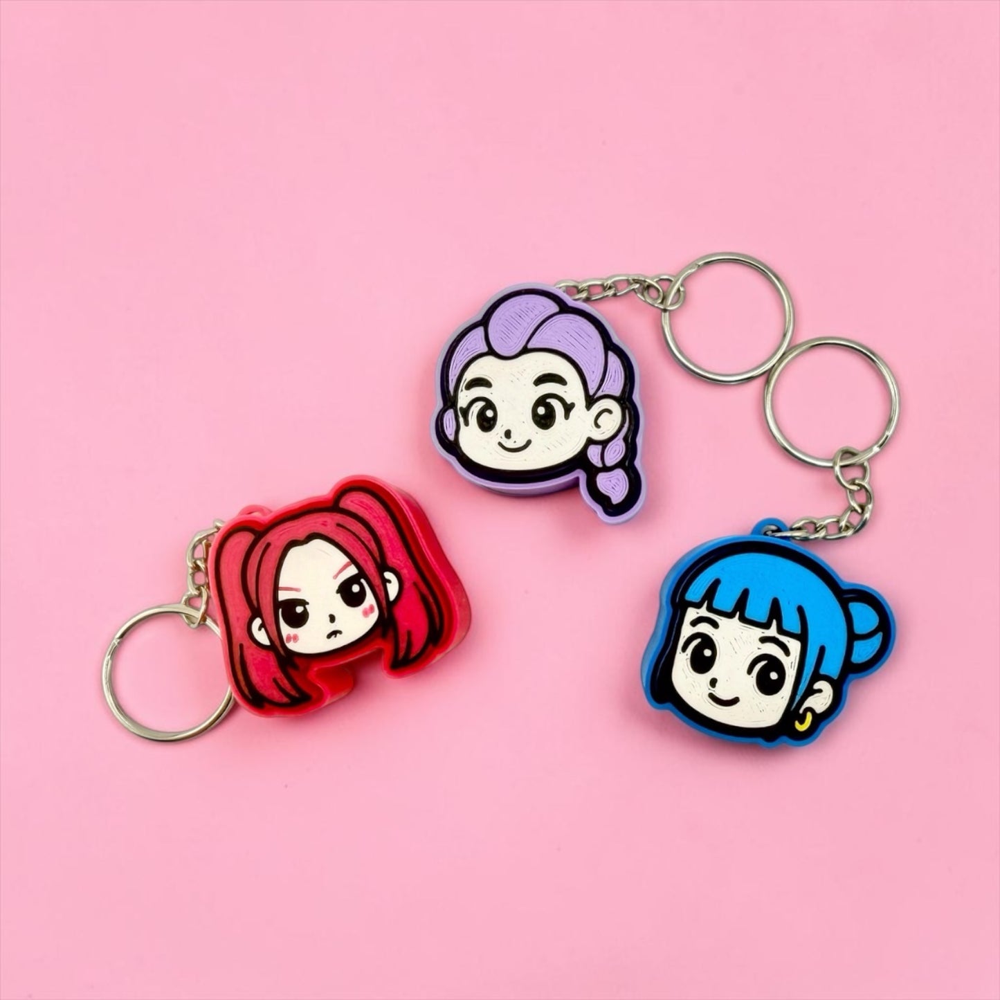 Three keychains with cartoon-style female characters on them, including a red character, a purple character, and a blue character, are displayed against a pink background.