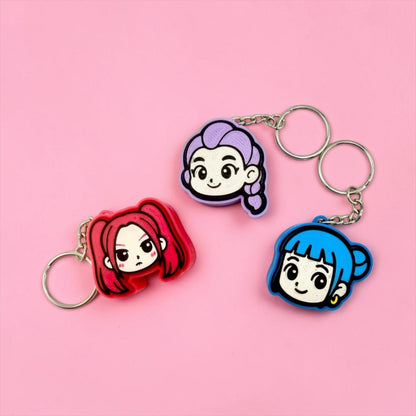 Three keychains with cartoon-style female characters on them, including a red character, a purple character, and a blue character, are displayed against a pink background.