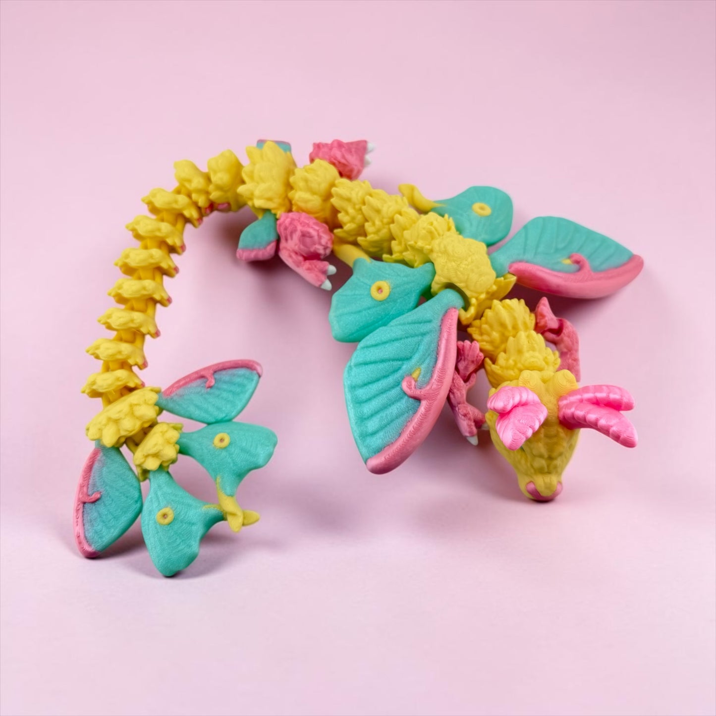 A colorful toy dragon made of plastic with yellow and pink scales, wings, and a pink body, sitting on a pink background.