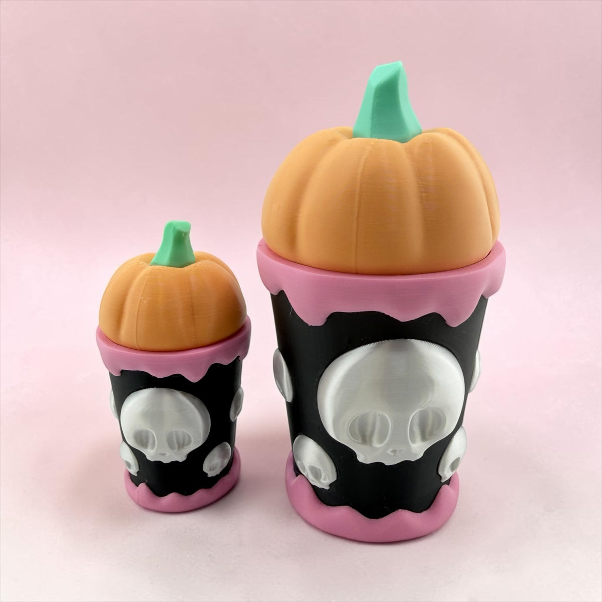 two small, colorful pumpkin-shaped containers with skull designs on them, set against a pink background.