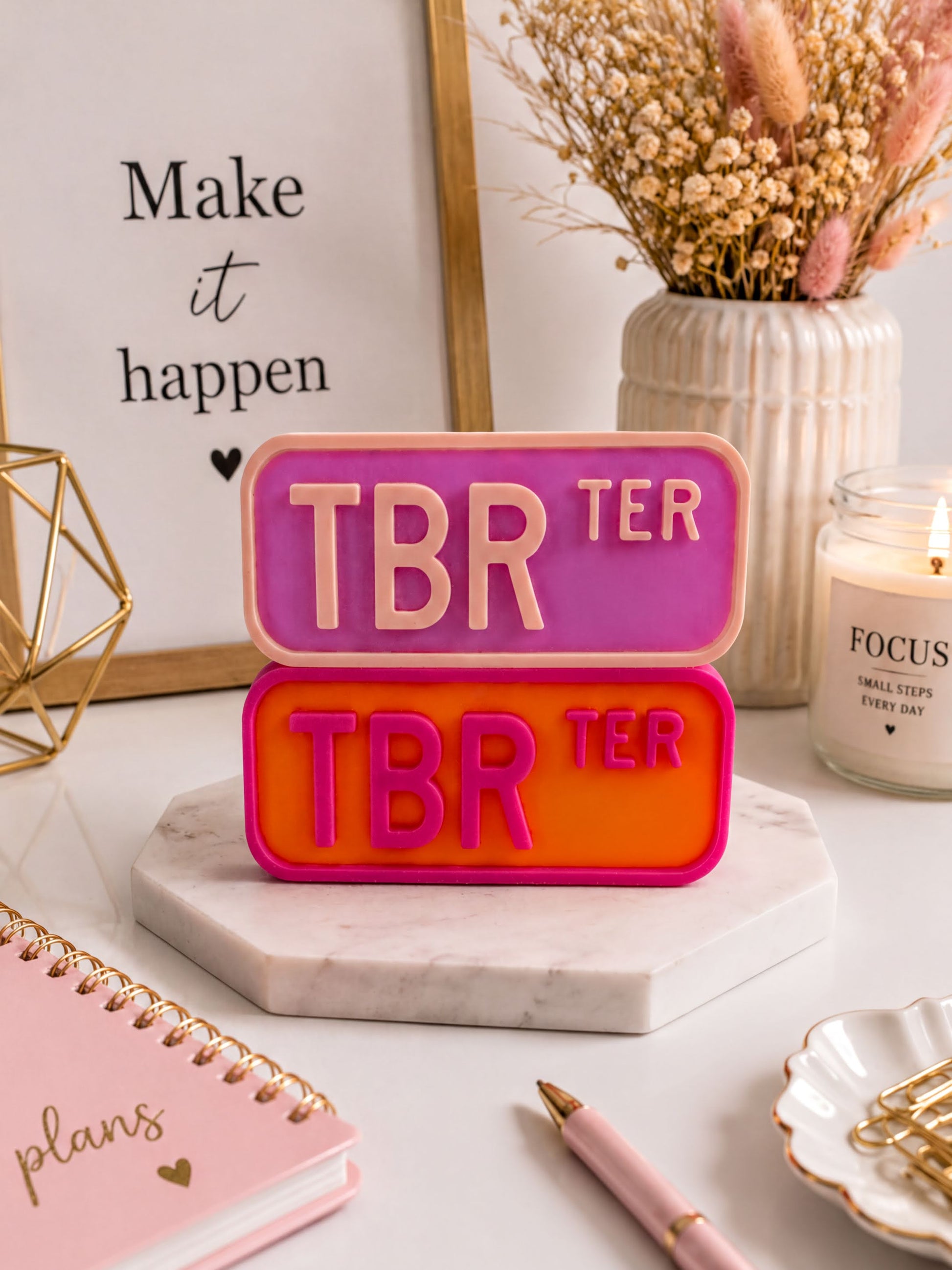 a desk with various items, including a pink notebook, a pink pen, a white vase with dried flowers, a framed quote, and a pink and orange object that says "TBR TER".