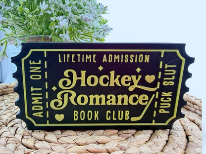 A black and yellow ticket-shaped sign with the text "Lifetime Admission Hockey Romance Book Club" on it, placed on a woven surface.