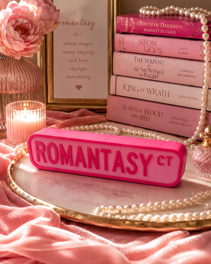a pink object with the word "ROMANTASY" on it, surrounded by a collection of books, pearls, and a vase with flowers.