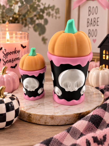 two small, orange-colored pumpkin-shaped containers with black and pink accents, placed on a marble surface. There are also small white pumpkins and a candle in the background, creating a festive Halloween atmosphere.