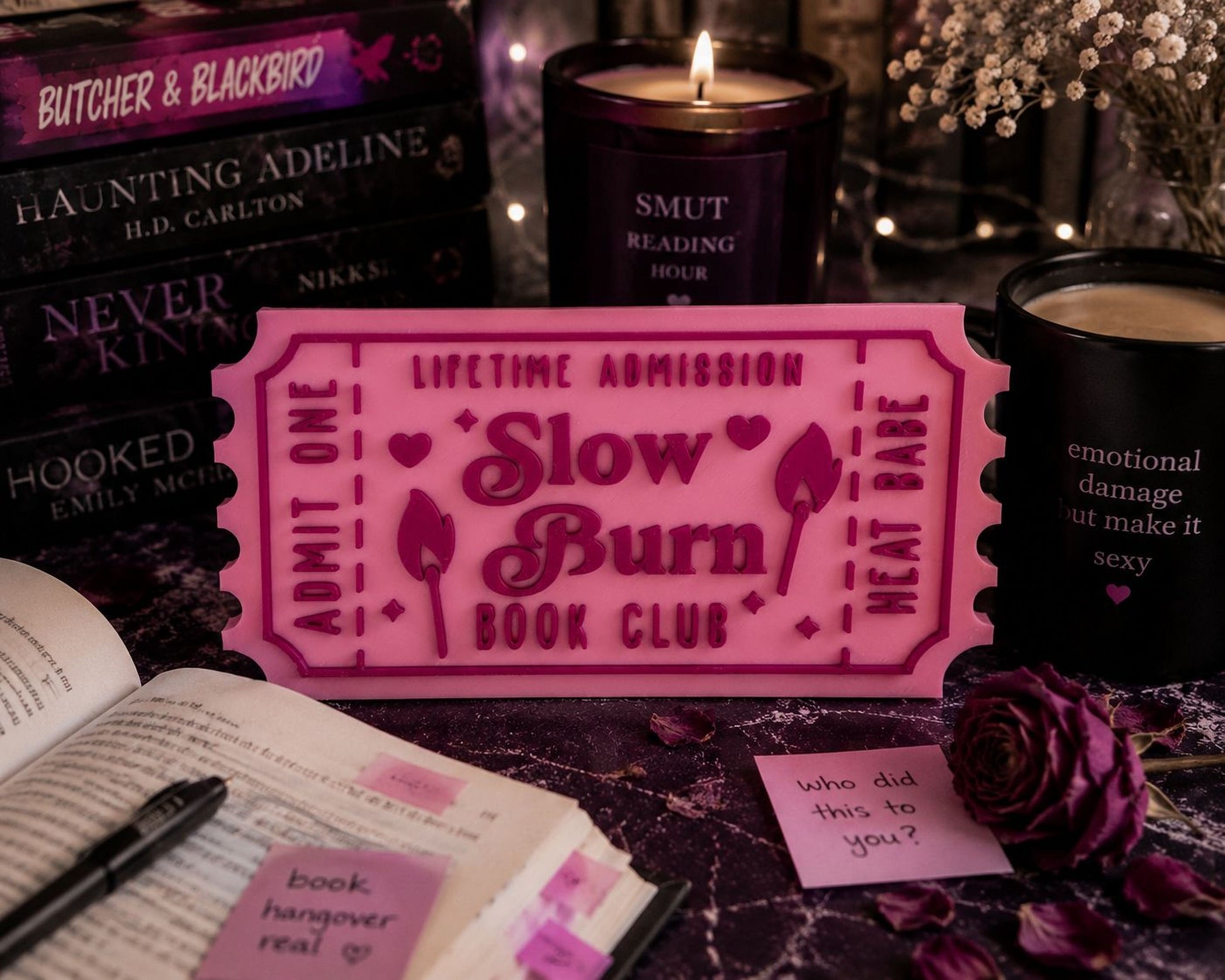 a pink ticket-shaped sign with the words "Lifetime Admission" and "Slow Burn Book Club" written on it, surrounded by various books, candles, and a rose.