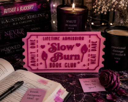 a pink ticket-shaped sign with the words "Lifetime Admission" and "Slow Burn Book Club" written on it, surrounded by various books, candles, and a rose.