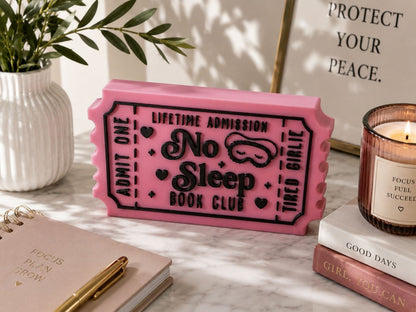 a pink ticket-shaped object with black text, placed on a marble surface next to a candle, a notebook, and a plant.