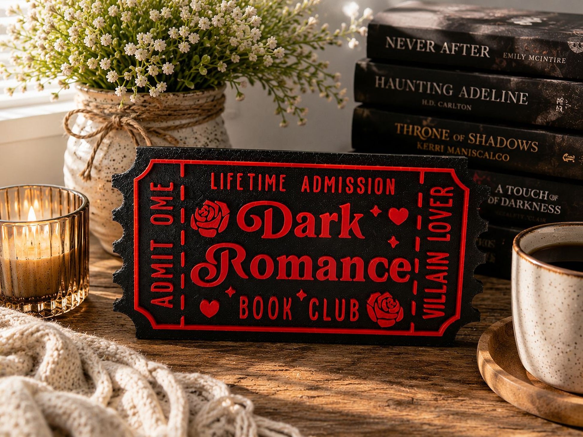 a book-themed sign with a red background and black text, surrounded by various items such as a vase of white flowers, a stack of books, and a cup of coffee.