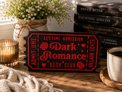a book-themed sign with a red background and black text, surrounded by various items such as a vase of white flowers, a stack of books, and a cup of coffee.