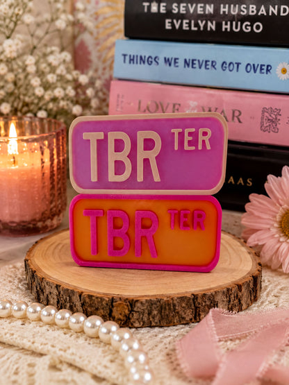 a pink and orange license plate with the text "TBR" on it, placed on a wooden surface surrounded by various decorative items such as pearls, a candle, and a bouquet of flowers.