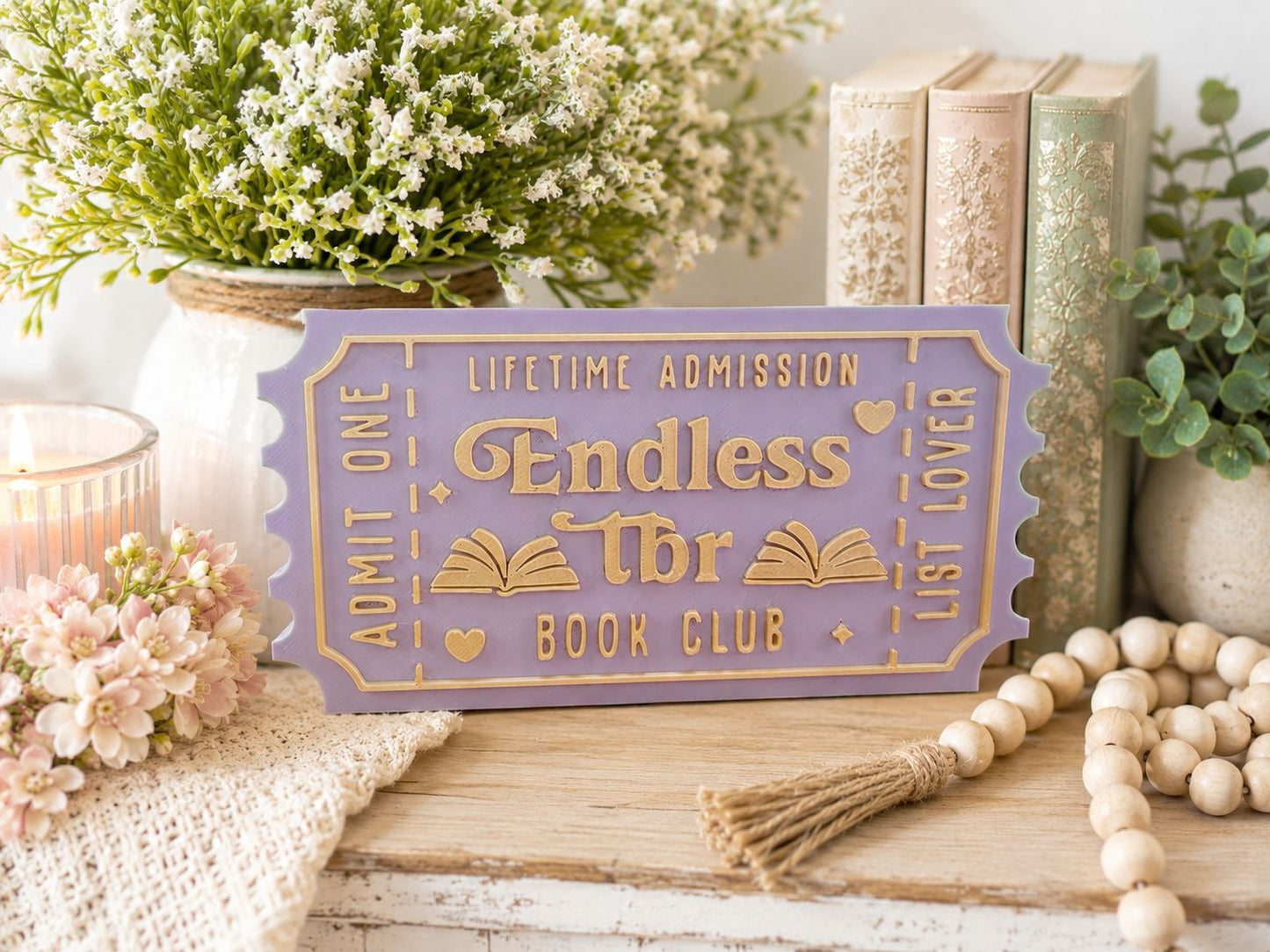 a purple ticket-shaped sign with gold lettering, surrounded by various decorative items such as a potted plant, books, and a beaded garland.