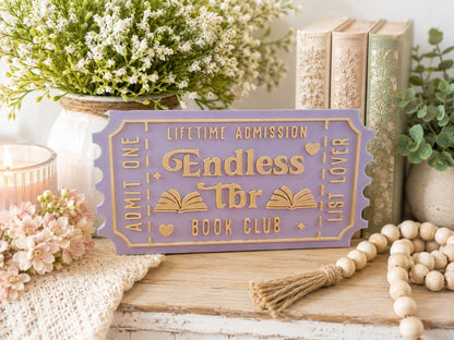 a purple ticket-shaped sign with gold lettering, surrounded by various decorative items such as a potted plant, books, and a beaded garland.