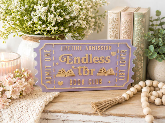 a purple ticket-shaped sign with gold lettering, surrounded by various decorative items such as a potted plant, books, and a beaded garland.