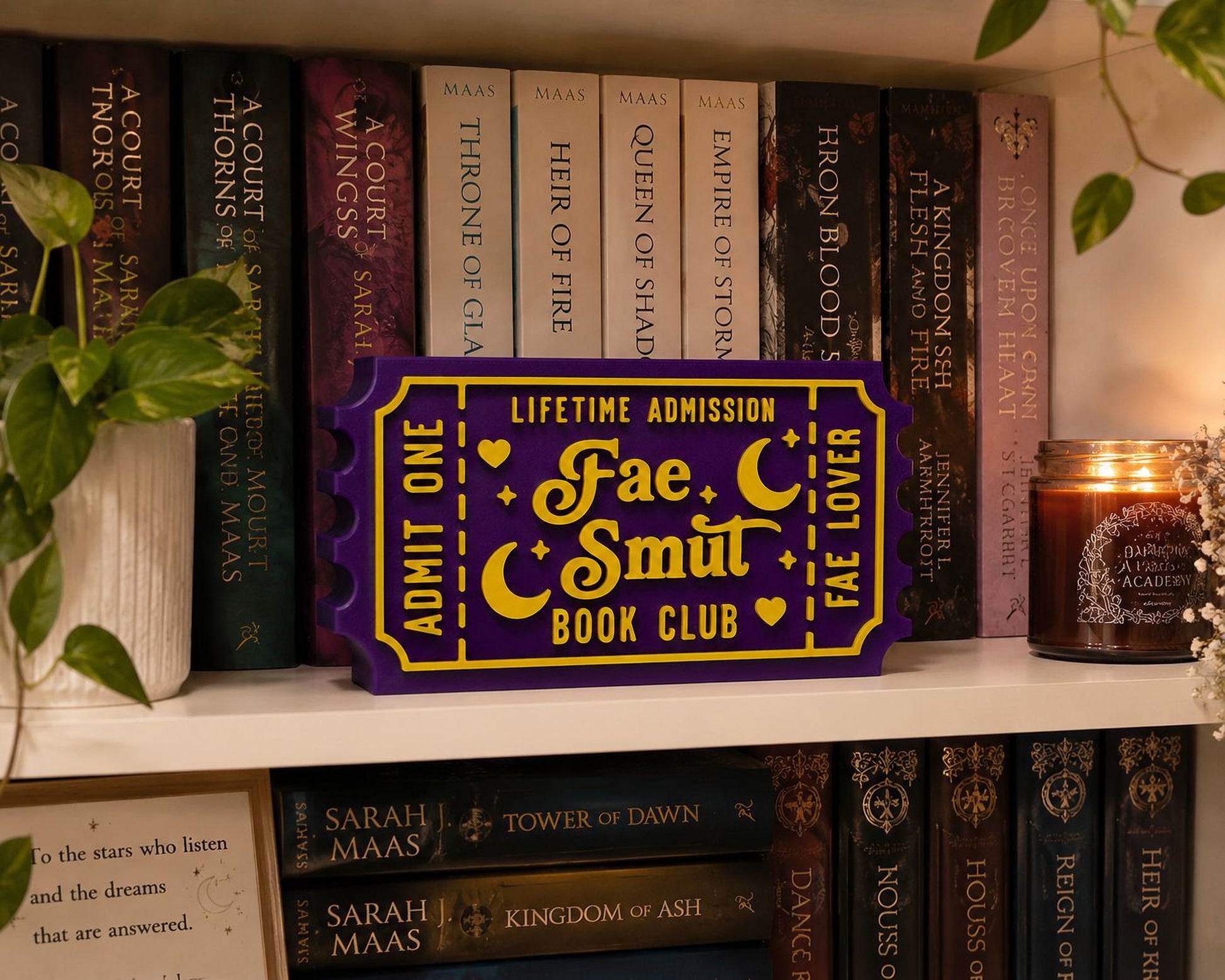 a purple ticket-shaped object with the text "Lifetime Admission Fae Smut Book Club" on it, placed on a bookshelf filled with various books.