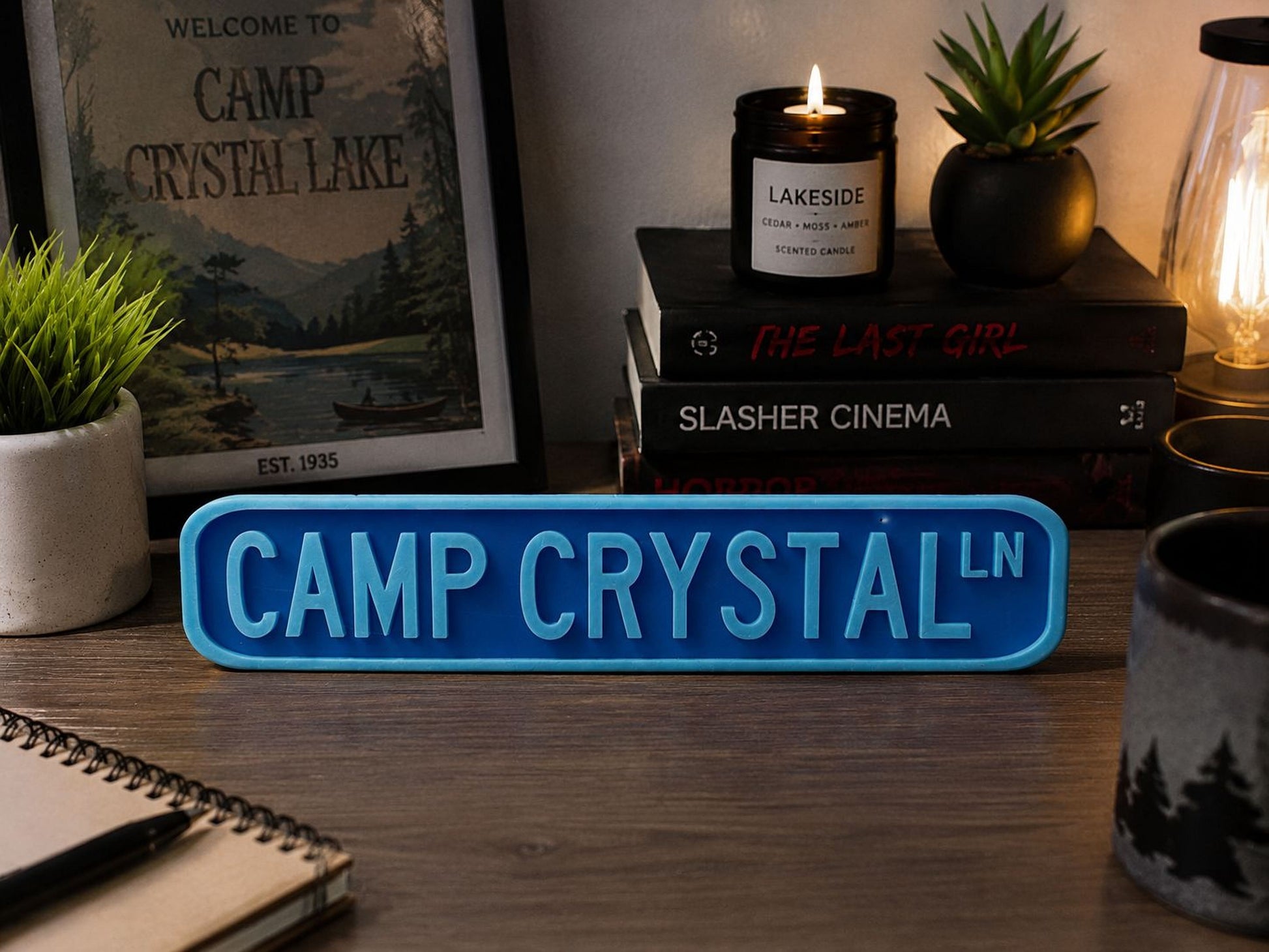 a desk with various items on it, including a blue sign that says "CAMP CRYSTAL LN", a candle, a mug, a notebook, and some books.