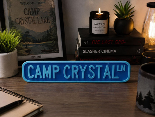 a desk with various items on it, including a blue sign that says "CAMP CRYSTAL LN", a candle, a mug, a notebook, and some books.