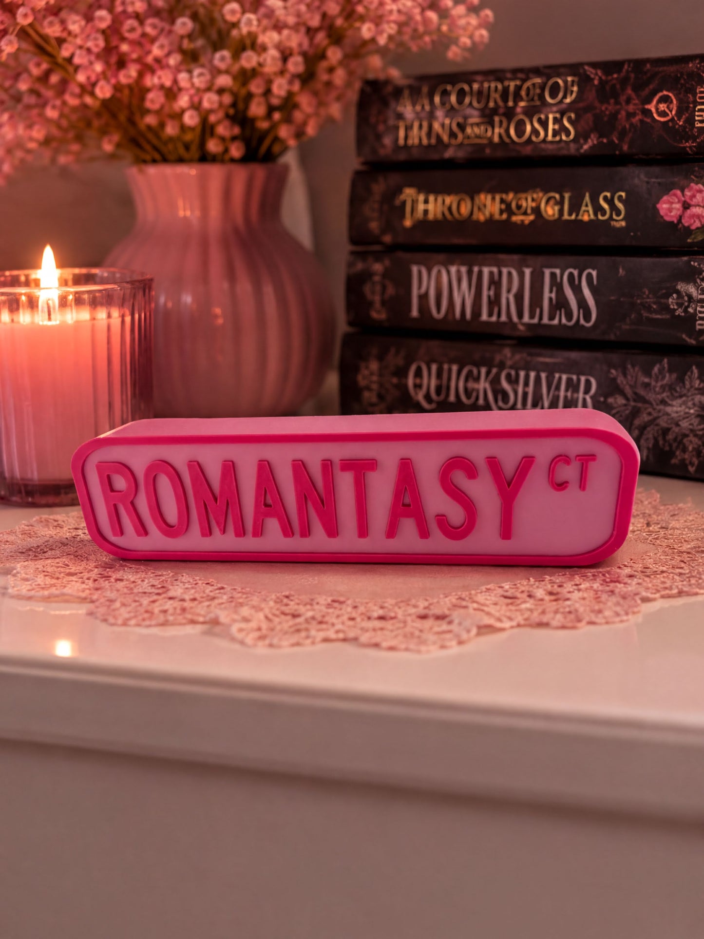 a pink rectangular object with the word "ROMANTASY" written on it, placed on a white surface. Behind the object, there are several books, including "A Court of Thrasher and Roses", "Throne of Glass", "Powerless", "Quicksilver", and "ROMANTASY". A lit candle is also visible in the background.