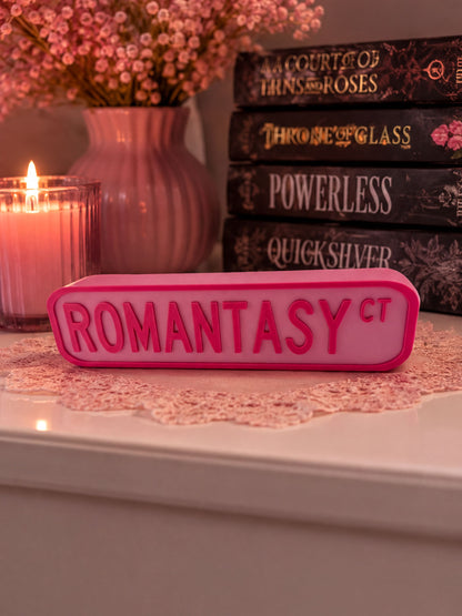 a pink rectangular object with the word "ROMANTASY" written on it, placed on a white surface. Behind the object, there are several books, including "A Court of Thrasher and Roses", "Throne of Glass", "Powerless", "Quicksilver", and "ROMANTASY". A lit candle is also visible in the background.