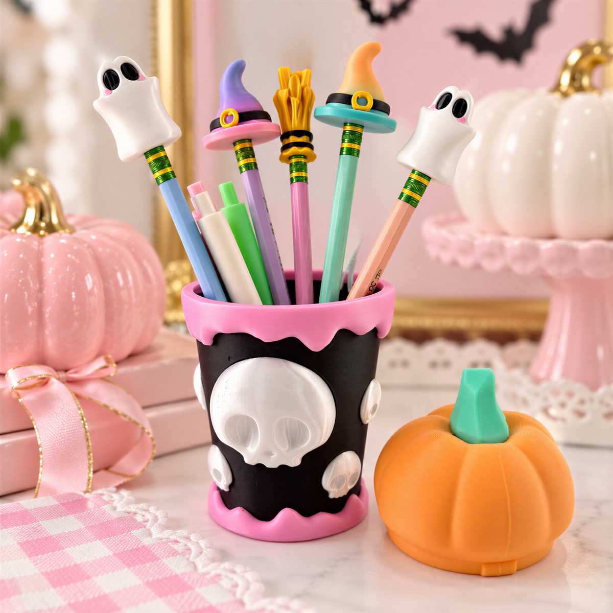 A collection of Halloween-themed items, including a cup filled with colorful pencils, a small orange pumpkin, and a pink pumpkin, are displayed on a table.