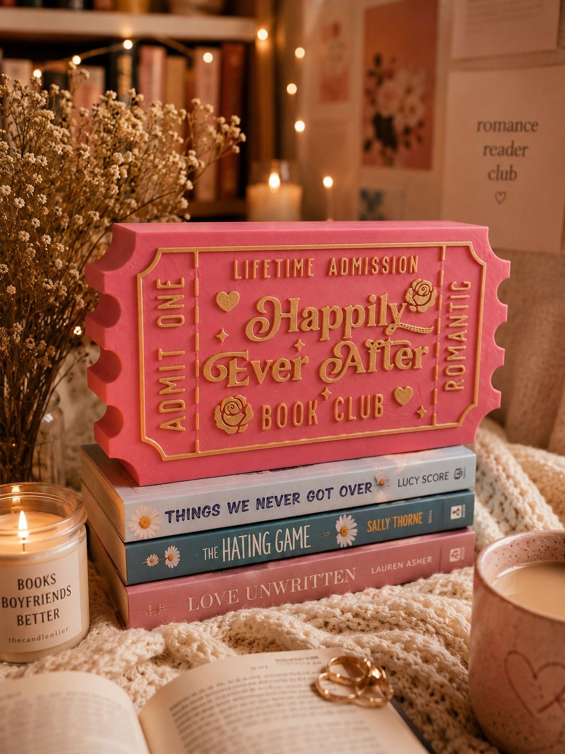 a pink ticket-shaped book club sign, a stack of books, a candle, and some decorative elements such as a vase and a heart-shaped object.