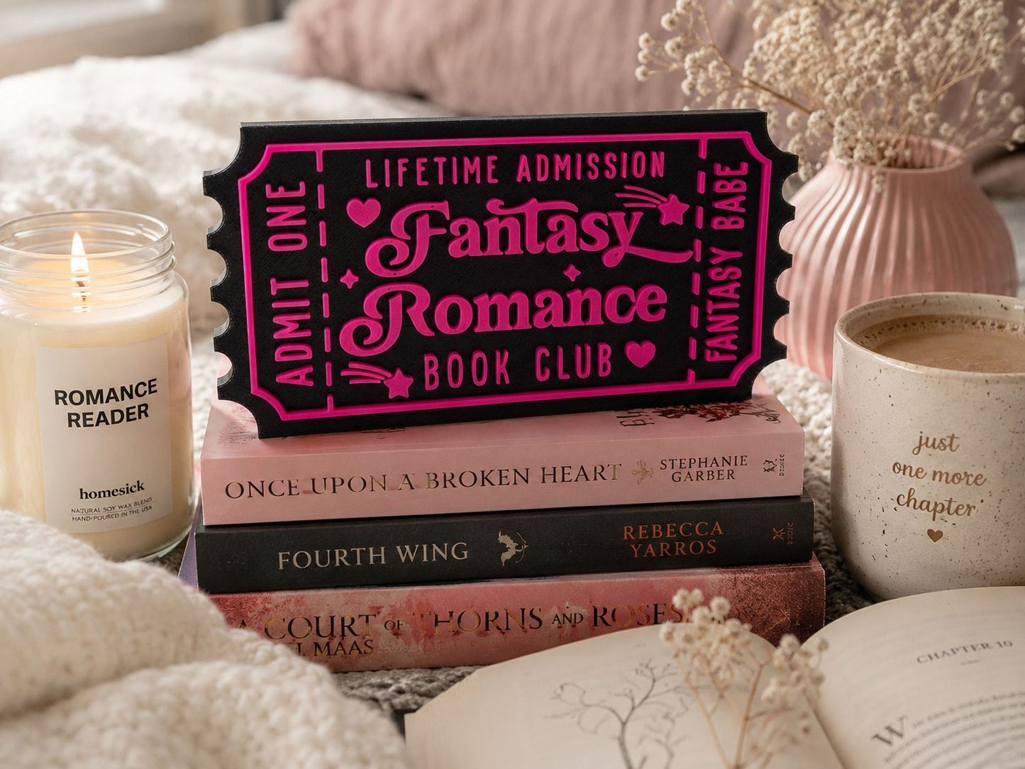 a stack of books, a pink ticket-shaped sign, a candle, and a mug, all arranged on a bed with a floral arrangement in the background.