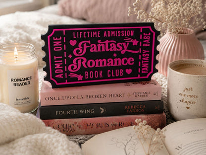 a stack of books, a pink ticket-shaped sign, a candle, and a mug, all arranged on a bed with a floral arrangement in the background.