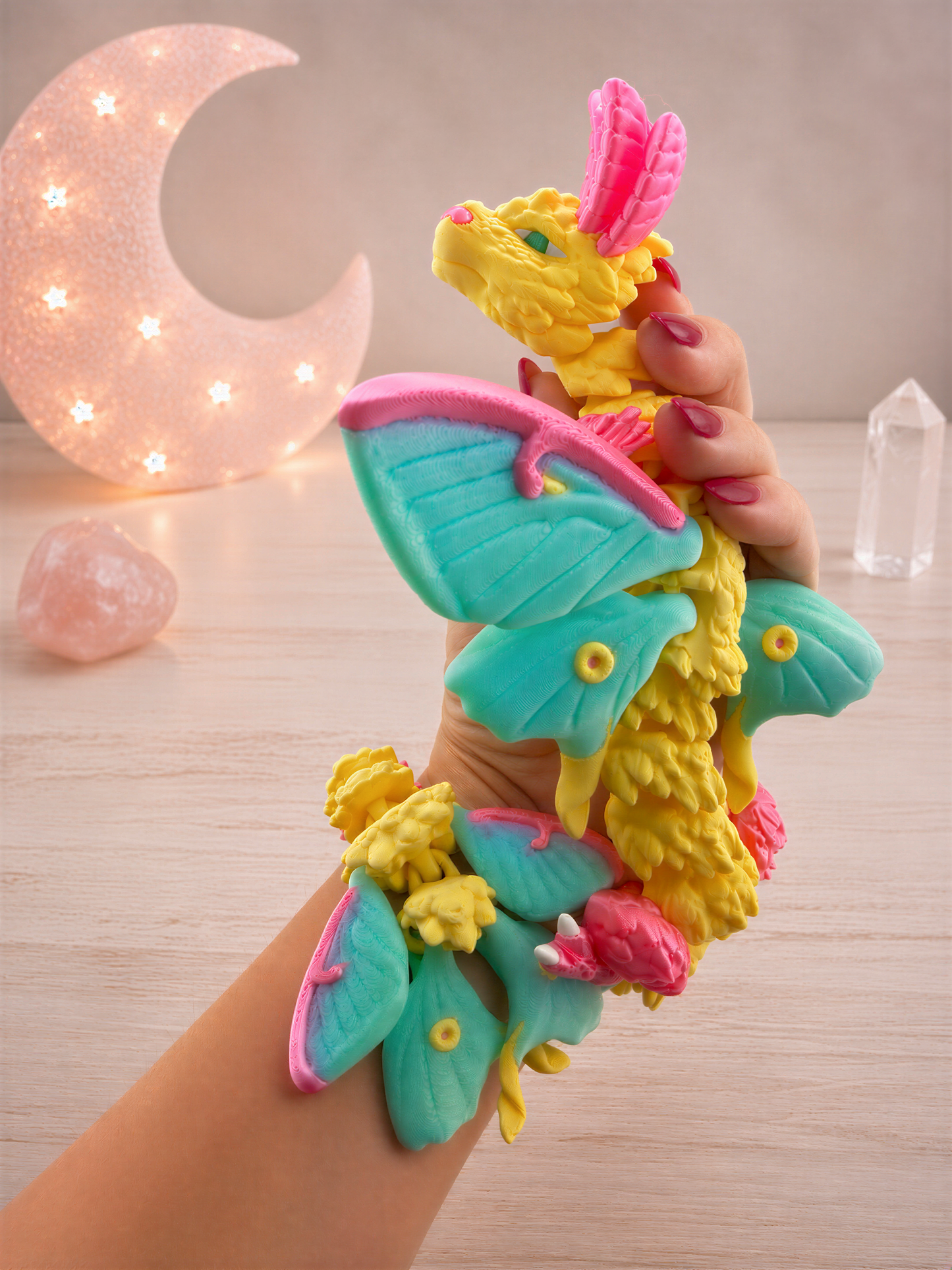 A hand holding a colorful, whimsical dragon-like toy with vibrant wings and a pink head, set against a backdrop of a crescent moon and crystals.