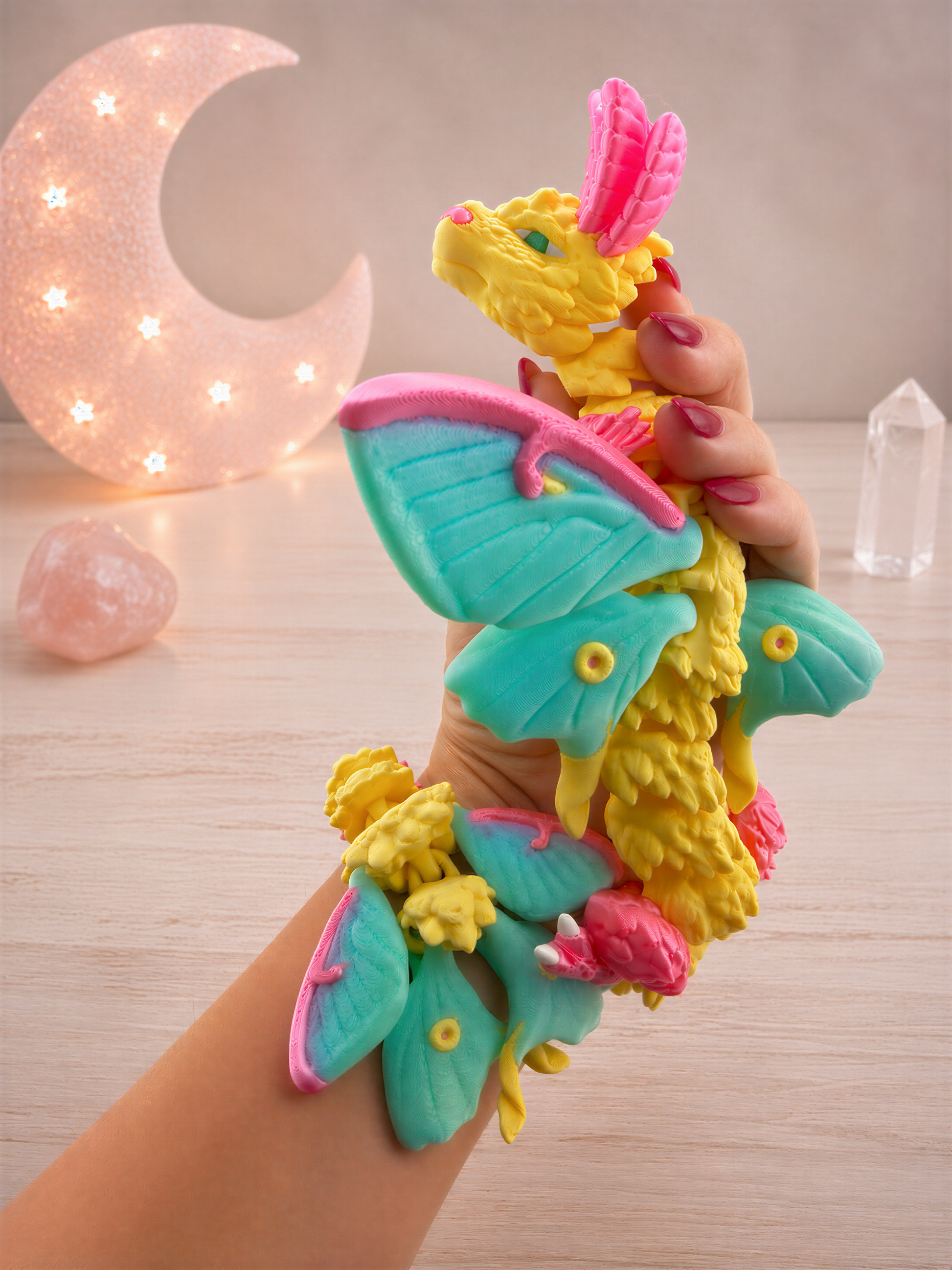A hand holding a colorful, whimsical dragon-like toy with vibrant wings and a pink head, set against a backdrop of a crescent moon and crystals.
