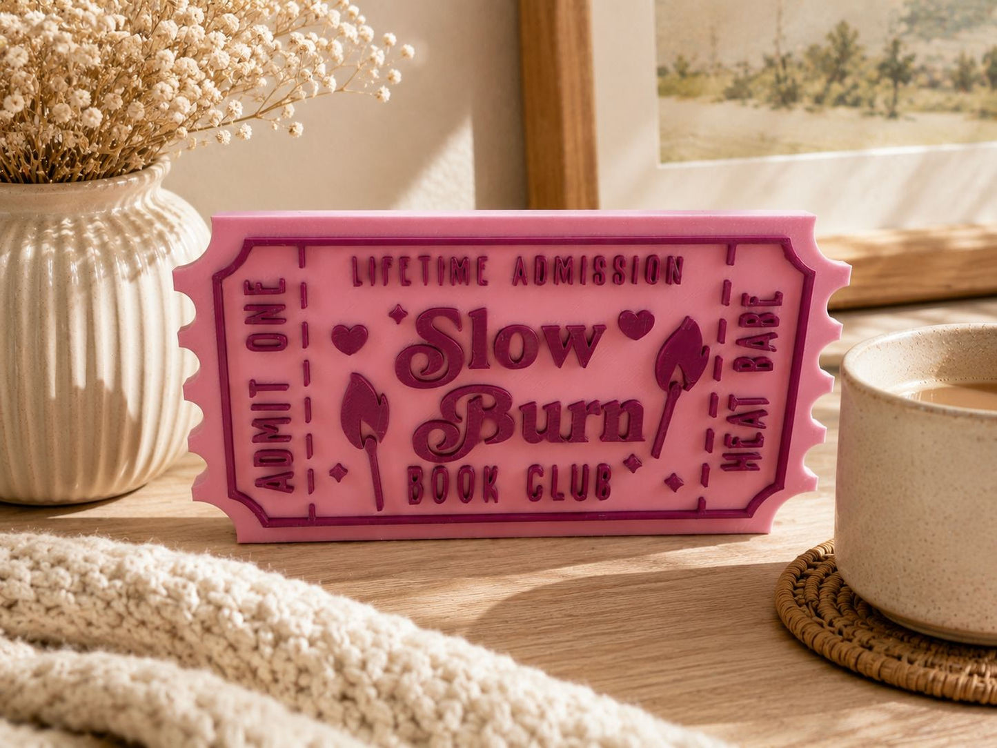 A pink ticket-shaped object with text on it, placed on a wooden surface next to a white mug and a beige vase with flowers.