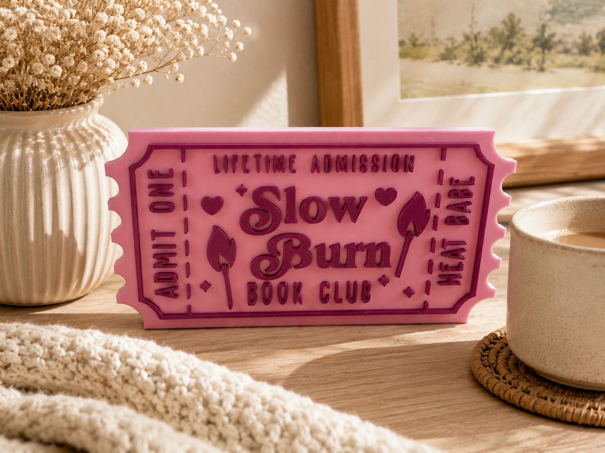 A pink ticket-shaped object with text on it, placed on a wooden surface next to a white mug and a beige vase with flowers.