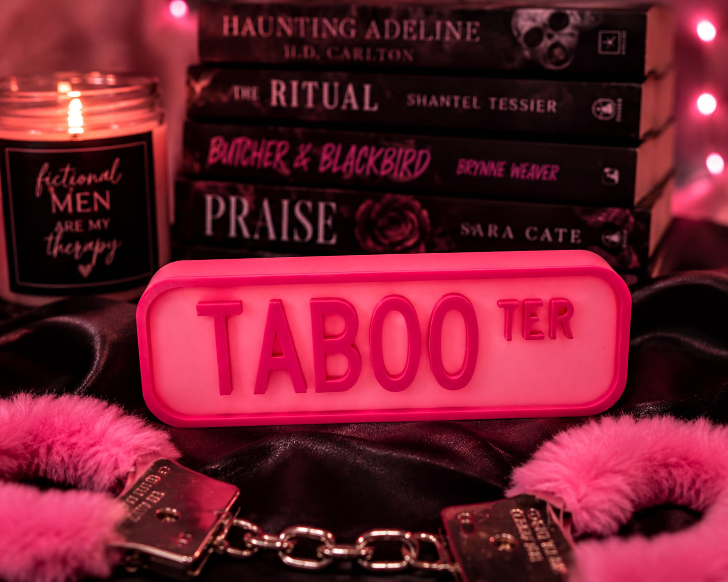 a pink sign with the word "TABOO" in large letters, surrounded by various books and accessories, including a candle, a pair of gloves, and a pair of handcuffs.