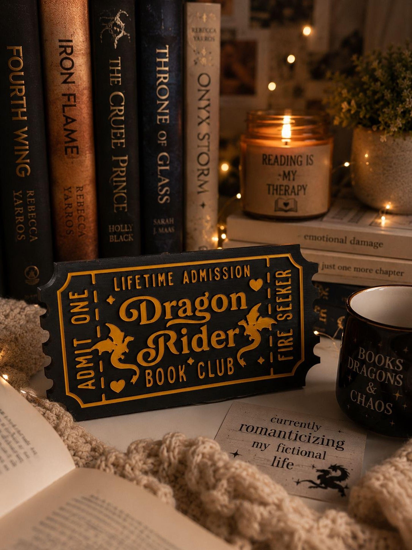 a cozy scene with a book titled "Dragon Rider Book Club" surrounded by other books, a candle, and a mug. The book cover displays the title "Dragon Rider" and the words "Lifetime Admission" and "Book Club". The candle reads "Reading is my therapy" and the mug says "Books Dragons Chaos". The background is decorated with twinkling lights, creating a warm and inviting atmosphere.