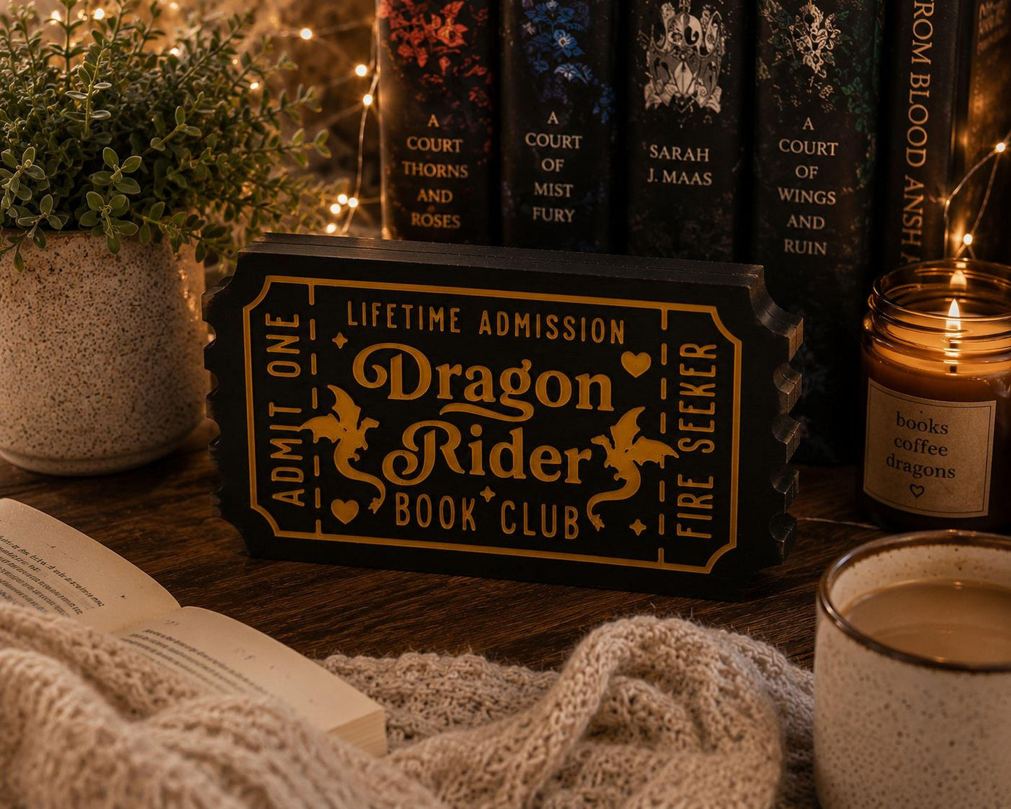 a cozy scene with a book titled "Dragon Rider Book Club" on a wooden surface, surrounded by a candle, a mug of coffee, and a knitted blanket.