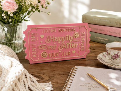 a pink ticket-shaped sign with the words "Happy Ever After" and "Lifetime Admission" on a wooden surface, surrounded by various items such as a teacup, a book, and a vase of flowers.