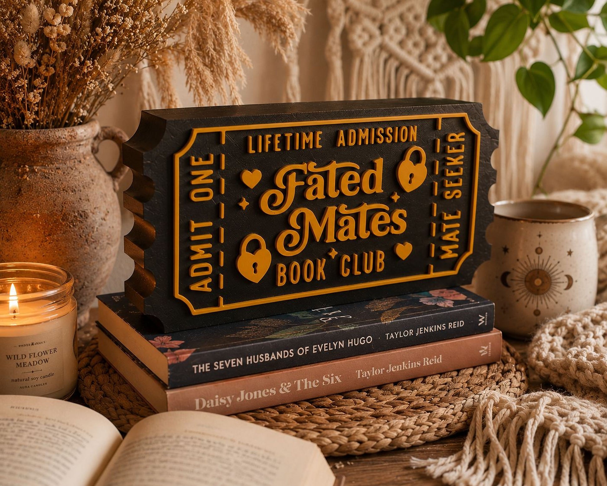 a stack of books, a candle, and a decorative ticket-shaped sign for a book club, all arranged on a woven mat.
