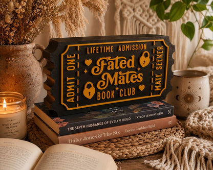 a stack of books, a candle, and a decorative ticket-shaped sign for a book club, all arranged on a woven mat.