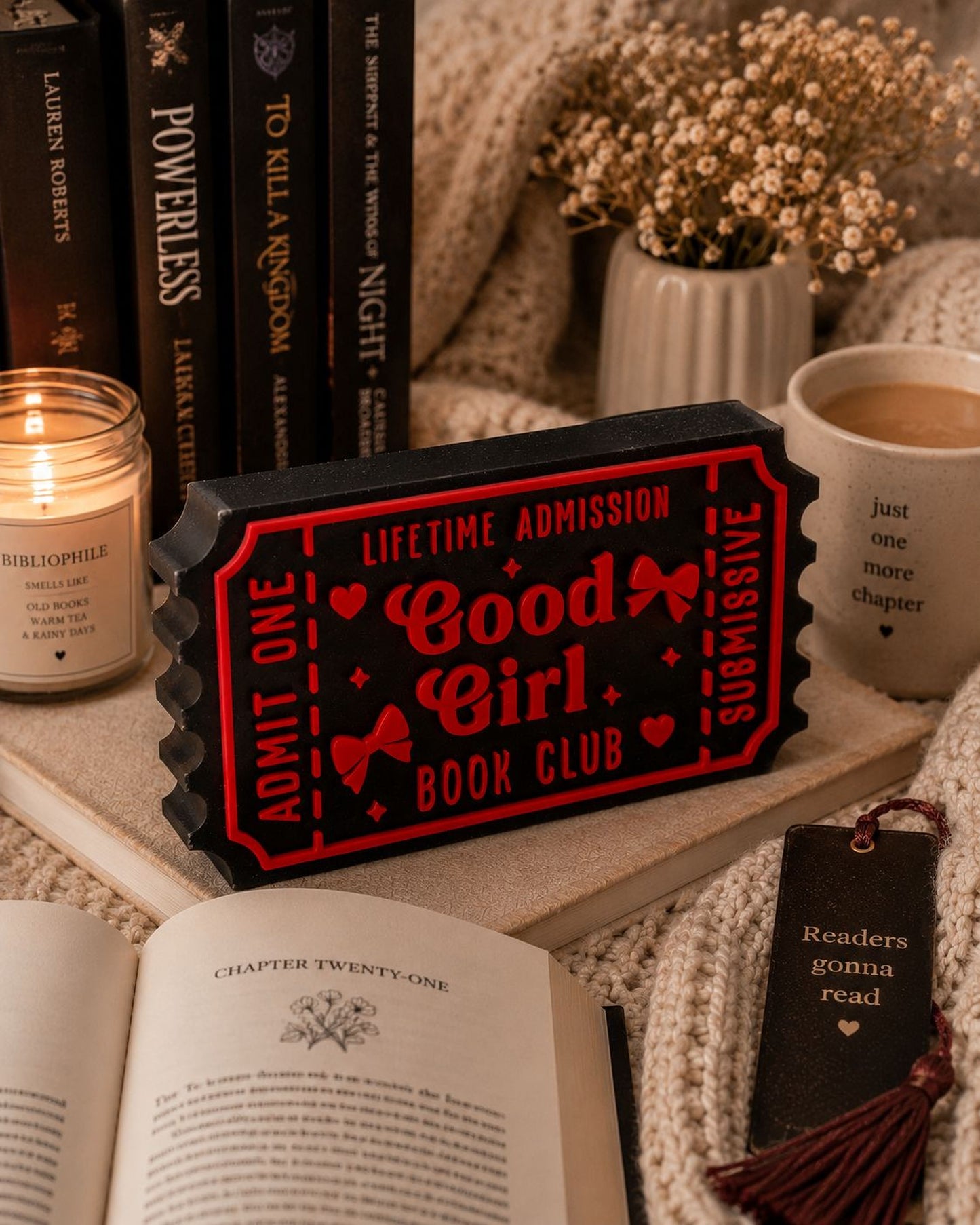 a cozy scene with a red ticket-shaped book club sign, a candle, a mug, and an open book on a bed.