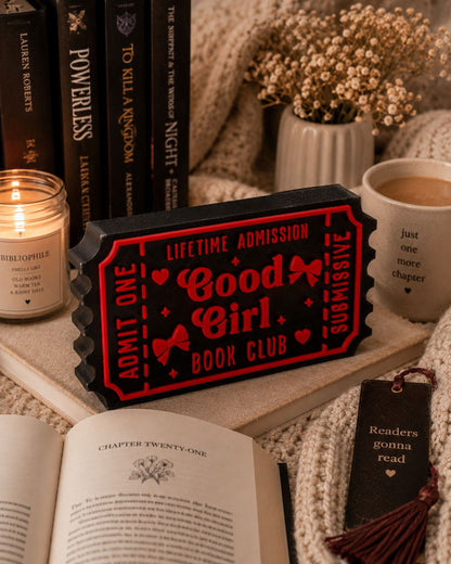 a cozy scene with a red ticket-shaped book club sign, a candle, a mug, and an open book on a bed.